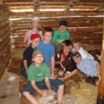 Eine Gruppe von Kindern sitzt und kniet auf Stroh in einer rustikalen Holzblockhütte, lächelt und posiert für die Kamera.