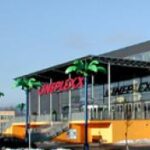 Modernes Gebäude mit großem "CINEPLEXX"-Schild, gläsernem Eingang und dekorativen Palmenstatuen davor; klarer Himmel im Hintergrund.