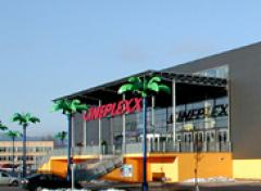 Modernes Gebäude mit großem "CINEPLEXX"-Schild, gläsernem Eingang und dekorativen Palmenstatuen davor; klarer Himmel im Hintergrund.