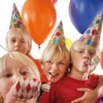 Vier Kinder mit Partyhüten und Krachmachern stehen dicht beieinander und lächeln, während im Hintergrund bunte Luftballons zu sehen sind.