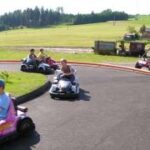 Mehrere Kinder fahren mit kleinen Gokarts auf einer Außenbahn mit Wiesen und Bäumen im Hintergrund.
