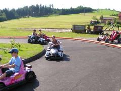 Mehrere Kinder fahren mit kleinen Gokarts auf einer Außenbahn mit Wiesen und Bäumen im Hintergrund.