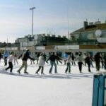 Eine Gruppe von Menschen, die tagsüber auf einer großen Eisbahn im Freien Schlittschuhlaufen, mit Gebäuden im Hintergrund.