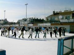 Eine Gruppe von Menschen, die tagsüber auf einer großen Eisbahn im Freien Schlittschuhlaufen, mit Gebäuden im Hintergrund.