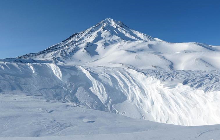 Schneebedeckter Berggipfel unter einem klaren blauen Himmel mit tiefen Schneeverwehungen und Graten im Vordergrund.