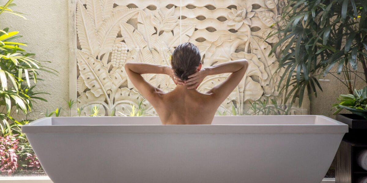 Eine Person mit nassem Haar sitzt in einer weißen Badewanne, mit Blick auf eine dekorative Wand mit tropischen Pflanzen, von hinten gesehen.
