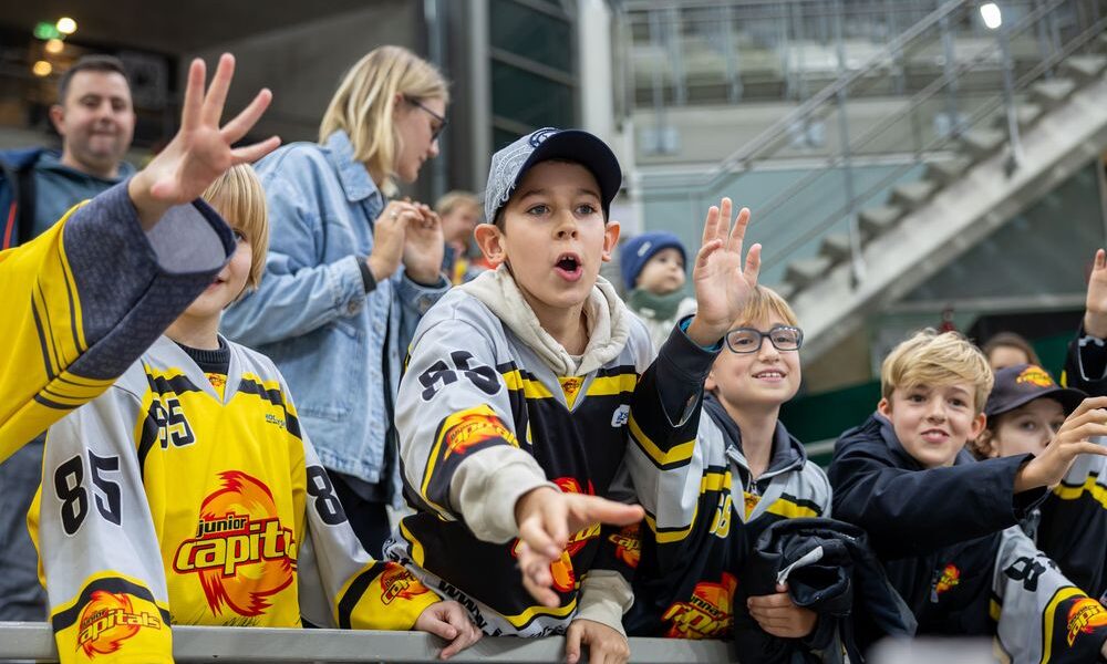 Eine Gruppe junger Eishockeyfans in Mannschaftstrikots reckt sich begeistert hinter einer Absperrung in einer Arena hervor, während im Hintergrund Zuschauer zu sehen sind.