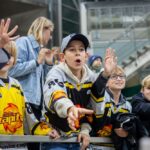 Eine Gruppe junger Eishockeyfans in Mannschaftstrikots reckt sich begeistert hinter einer Absperrung in einer Arena hervor, während im Hintergrund Zuschauer zu sehen sind.