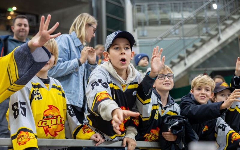Eine Gruppe junger Eishockeyfans in Mannschaftstrikots reckt sich begeistert hinter einer Absperrung in einer Arena hervor, während im Hintergrund Zuschauer zu sehen sind.