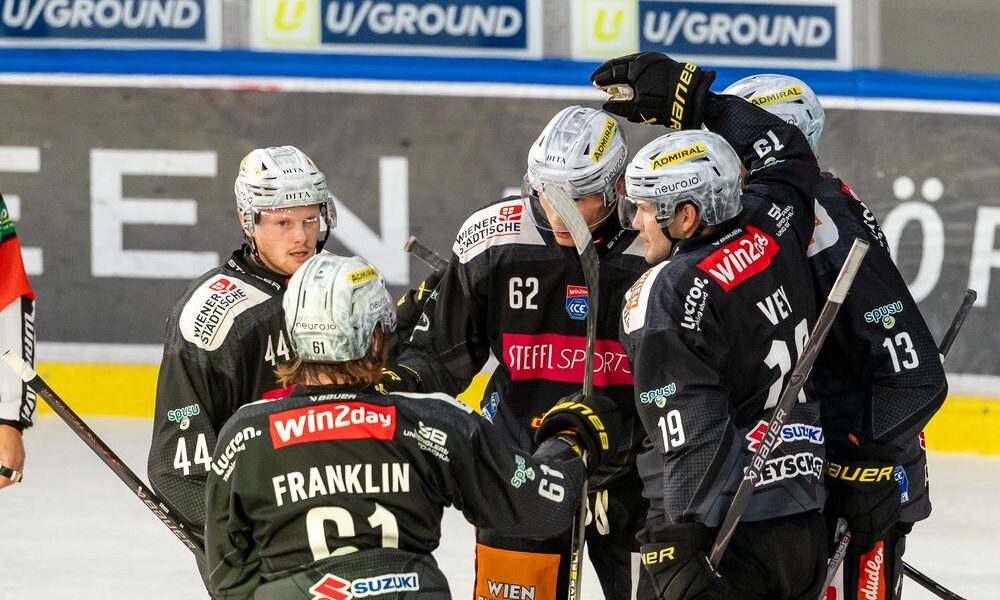 Eishockeyspieler in schwarzen Trikots versammeln sich auf dem Eis und scheinen während eines Spiels zu feiern, während im Hintergrund Werbung auf den Banden zu sehen ist.