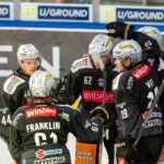 Eishockeyspieler in schwarzen Trikots versammeln sich auf dem Eis und scheinen während eines Spiels zu feiern, während im Hintergrund Werbung auf den Banden zu sehen ist.