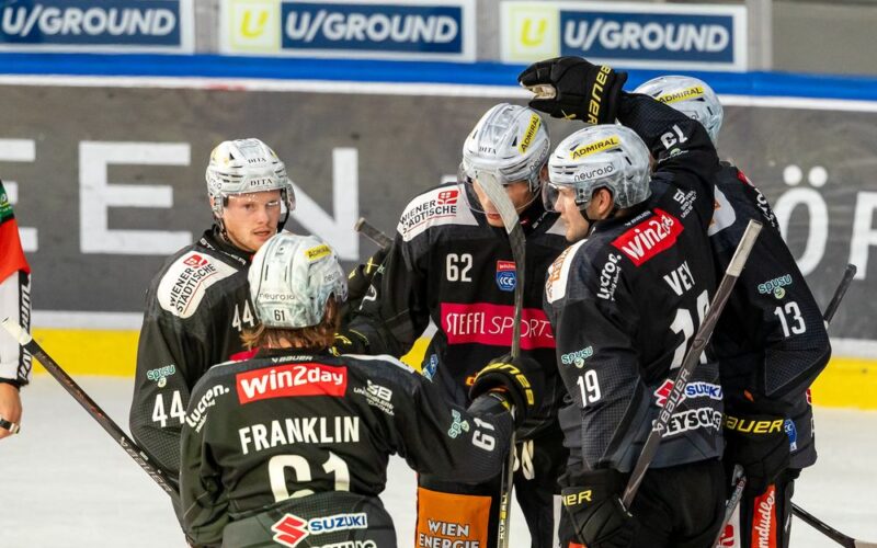 Eishockeyspieler in schwarzen Trikots versammeln sich auf dem Eis und scheinen während eines Spiels zu feiern, während im Hintergrund Werbung auf den Banden zu sehen ist.