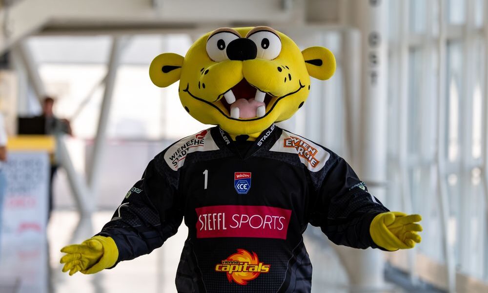 Eine Person in einem gelben Raubkatzenkostüm, die ein schwarzes Trikot mit den Logos "Steel Sports" und "Capitals" trägt, steht mit leicht ausgestreckten Armen in der Halle.
