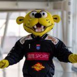 Eine Person in einem gelben Raubkatzenkostüm, die ein schwarzes Trikot mit den Logos "Steel Sports" und "Capitals" trägt, steht mit leicht ausgestreckten Armen in der Halle.