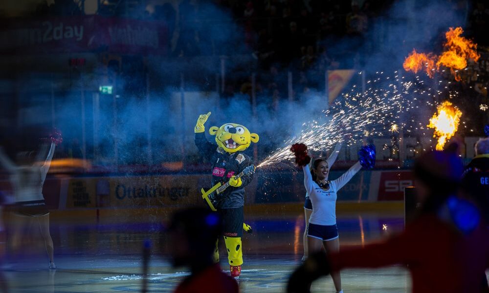 Ein Sportmaskottchen in einem gelben Kostüm steht auf einer Eisbahn mit einer Gitarrenrequisite in der Hand, umgeben von Cheerleadern und pyrotechnischen Effekten während einer Vorstellung.