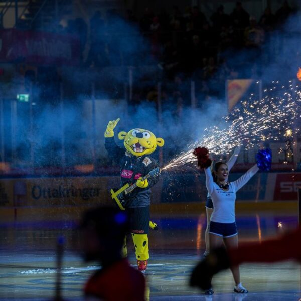 Ein Sportmaskottchen in einem gelben Kostüm steht auf einer Eisbahn mit einer Gitarrenrequisite in der Hand, umgeben von Cheerleadern und pyrotechnischen Effekten während einer Vorstellung.