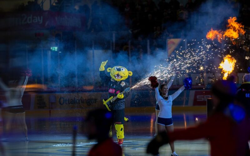 Ein Sportmaskottchen in einem gelben Kostüm steht auf einer Eisbahn mit einer Gitarrenrequisite in der Hand, umgeben von Cheerleadern und pyrotechnischen Effekten während einer Vorstellung.