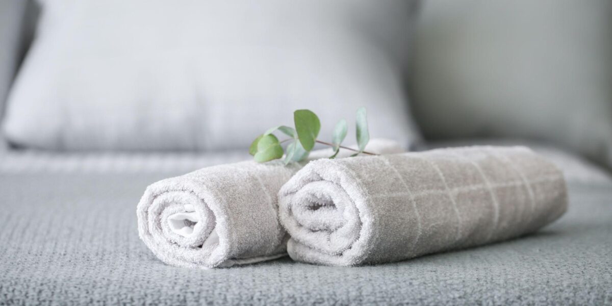 Zwei aufgerollte graue Handtücher mit einem kleinen grünen Zweig liegen auf einem ordentlich gemachten grauen Bett mit Kissen im Hintergrund.