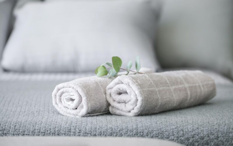 Zwei aufgerollte graue Handtücher mit einem kleinen grünen Zweig liegen auf einem ordentlich gemachten grauen Bett mit Kissen im Hintergrund.
