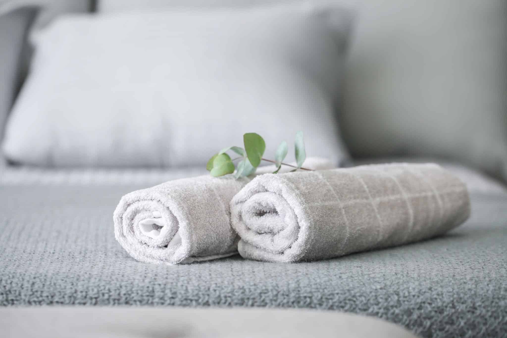Zwei aufgerollte graue Handtücher mit einem kleinen grünen Zweig liegen auf einem ordentlich gemachten grauen Bett mit Kissen im Hintergrund.