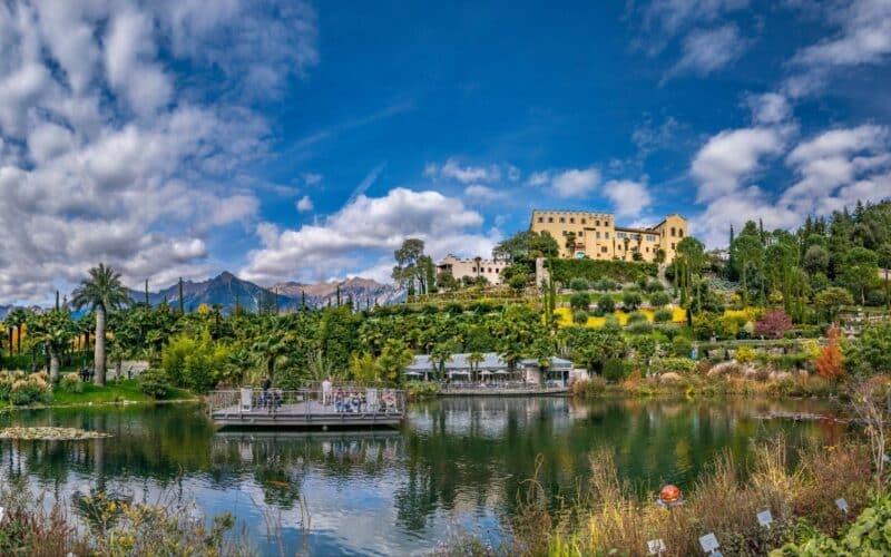 Ein terrassenförmig angelegter botanischer Garten mit einem großen Teich, üppigem Grün und einer gelben Villa auf einem Hügel vor der Kulisse von Bergen und einem teilweise bewölkten Himmel.