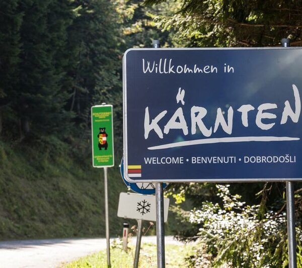 Ein blaues Straßenschild mit der Aufschrift "Willkommen in Kärnten" und darunter "Welcome, Benvenuti, Dobrodošli" markiert den Eingang zur Region Kärnten in Österreich. Im Hintergrund sind Wald und andere Schilder zu sehen.