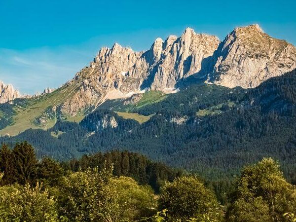 Schroffe, felsige Berggipfel erheben sich über bewaldete Hügel und grüne Vegetation unter einem klaren blauen Himmel.