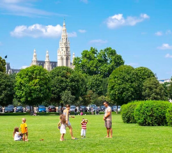 Erwachsene und Kinder stehen und spielen auf einer Wiese mit Bäumen, geparkten Autos und einem großen gotischen Gebäude mit Türmen im Hintergrund unter einem blauen Himmel.