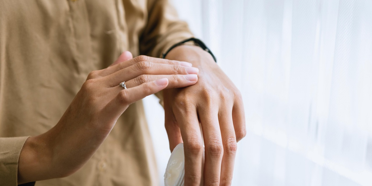 Eine Person cremt sich in der Nähe eines Fensters den Handrücken ein, trägt ein hellbraunes Hemd und einen Ring an einem Finger.