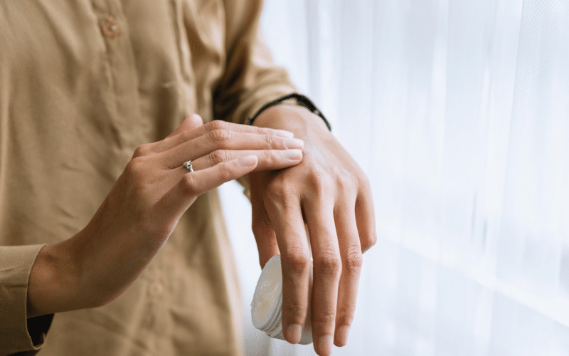 Eine Person cremt sich in der Nähe eines Fensters den Handrücken ein, trägt ein hellbraunes Hemd und einen Ring an einem Finger.
