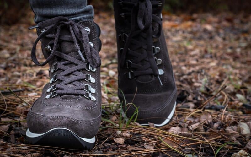 Nahaufnahme einer Person in dunklen Wanderstiefeln, die auf einem mit getrockneten Blättern und Tannennadeln bedeckten Waldboden steht.