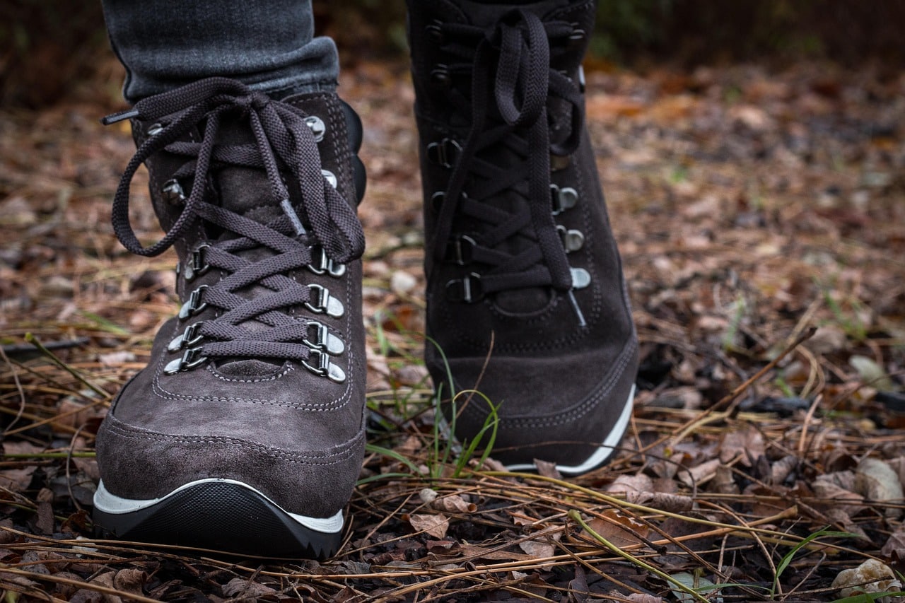 Nahaufnahme einer Person in dunklen Wanderstiefeln, die auf einem mit getrockneten Blättern und Tannennadeln bedeckten Waldboden steht.