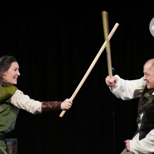 Zwei Schauspieler auf der Bühne, die einen Schwertkampf mit Holzstöcken vortäuschen, beide in mittelalterlichen Kostümen, vor einem schwarzen Hintergrund.