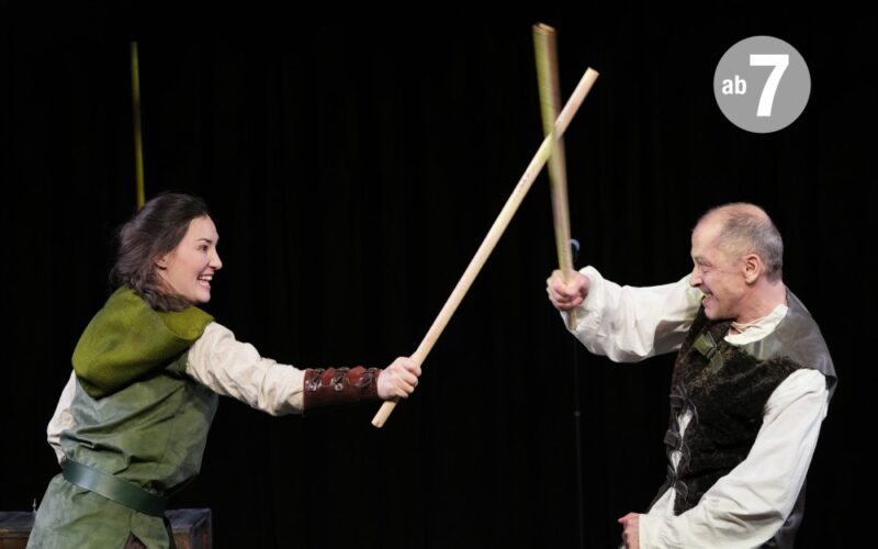 Zwei Schauspieler auf der Bühne, die einen Schwertkampf mit Holzstöcken vortäuschen, beide in mittelalterlichen Kostümen, vor einem schwarzen Hintergrund.