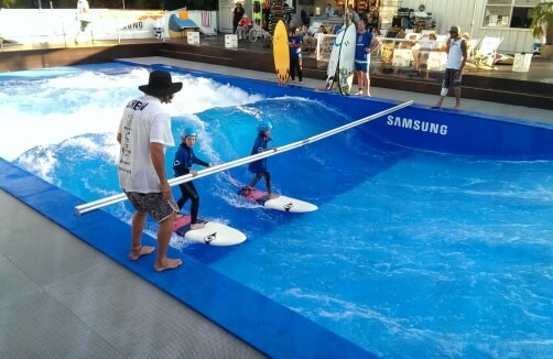 Drei Personen üben das Surfen in einem künstlichen Wellenbad, während ein Lehrer sie mit einer Metallstange anleitet. Andere Personen schauen vom Deck im Hintergrund zu.