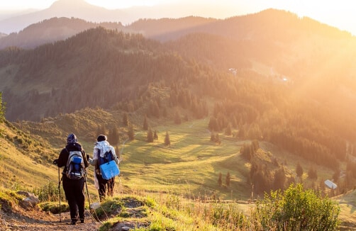 Zwei Wanderer mit Rucksäcken gehen bei Sonnenaufgang auf einem grasbewachsenen Berghang entlang, umgeben von bewaldeten Hügeln und fernen Gipfeln.