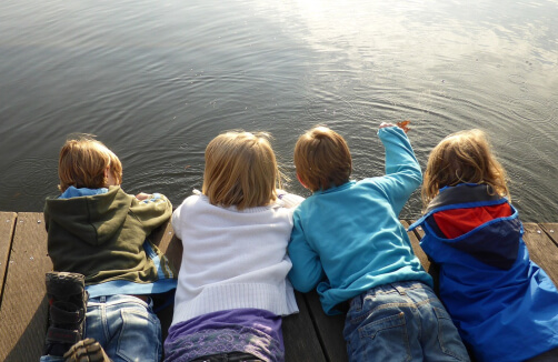 Vier Kinder liegen auf dem Bauch auf einem Holzsteg und schauen auf die Wasseroberfläche.