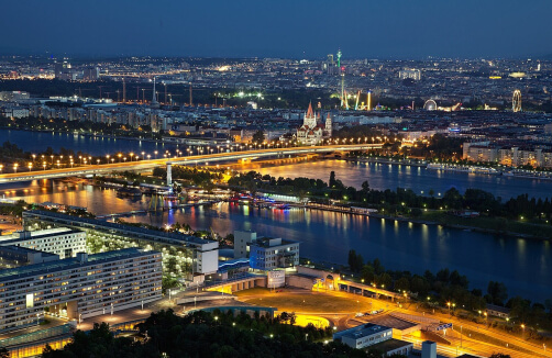 Eine Stadtlandschaft in der Abenddämmerung mit beleuchteten Brücken über einen Fluss, Gebäuden im Vordergrund und einem Riesenrad und einer Kirche in der Ferne.