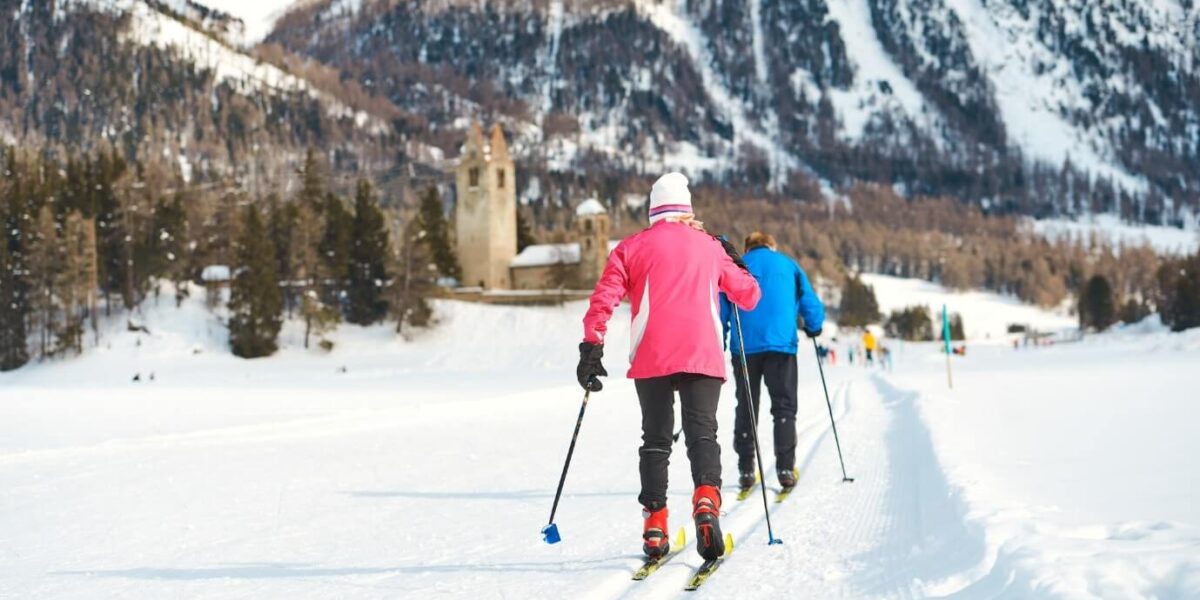 Zwei Personen beim Skilanglauf auf einer verschneiten Loipe, umgeben von schneebedeckten Bergen und Bäumen, mit einem alten Steingebäude im Hintergrund.