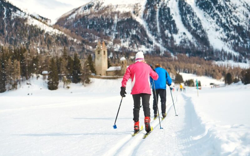 Zwei Personen beim Skilanglauf auf einer verschneiten Loipe, umgeben von schneebedeckten Bergen und Bäumen, mit einem alten Steingebäude im Hintergrund.