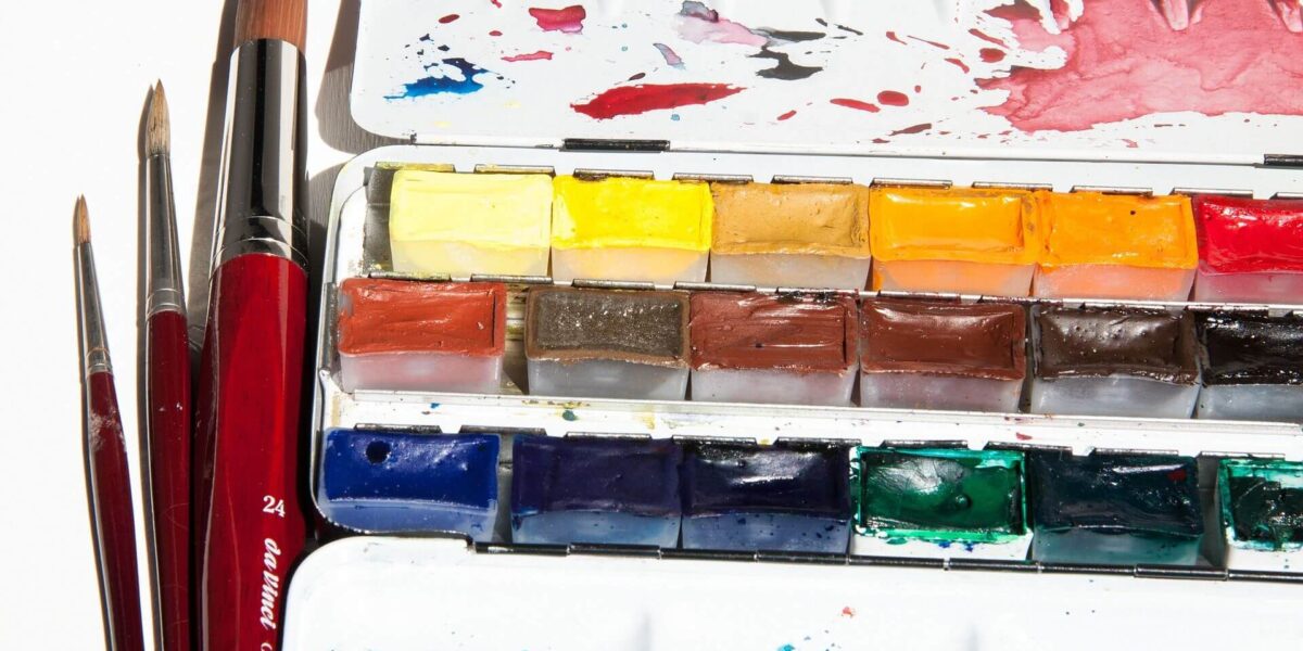 Drei Pinsel liegen neben einer offenen Aquarellpalette, die verschiedene bunte Farbblöcke enthält, mit Flecken auf dem Deckel und der Mischschale.