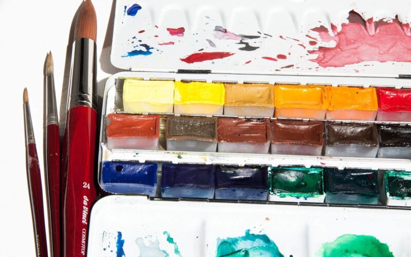 Drei Pinsel liegen neben einer offenen Aquarellpalette, die verschiedene bunte Farbblöcke enthält, mit Flecken auf dem Deckel und der Mischschale.