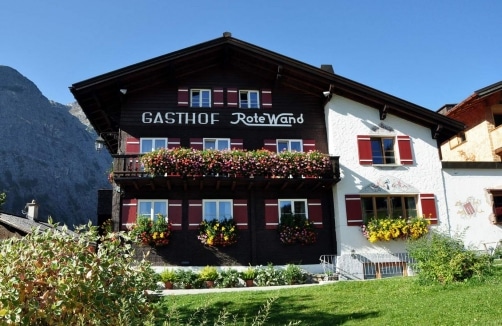 Ein traditioneller Alpengasthof mit Blumenkästen an Fenstern und Balkonen, mit dem Schild "Gasthof Rote Wand" vor einem strahlend blauen Himmel.