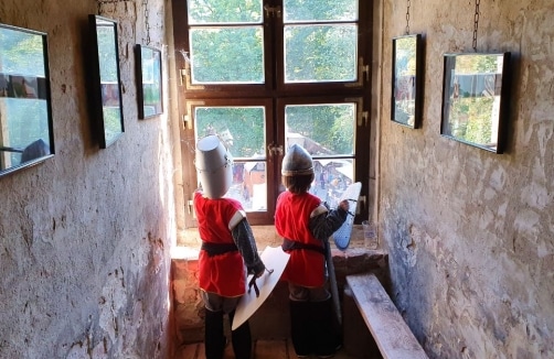 Zwei Kinder, die als mittelalterliche Ritter gekleidet sind, stehen an einem Fenster in einem Steingebäude und schauen nach draußen. Um sie herum hängen gerahmte Bilder an den rauen Wänden.