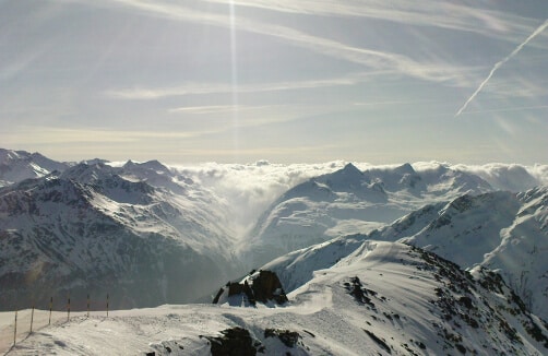 Schneebedeckte Gebirgskette unter einem teilweise bewölkten Himmel mit Sonnenstrahlen und Kondensstreifen; die Gipfel erheben sich über die Wolken im Hintergrund.