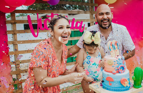 Drei Personen feiern auf einer Geburtstagsparty mit Konfetti, einer bunten Torte und Dekorationen; ein Kind steht zwischen zwei Erwachsenen, die alle lächeln.