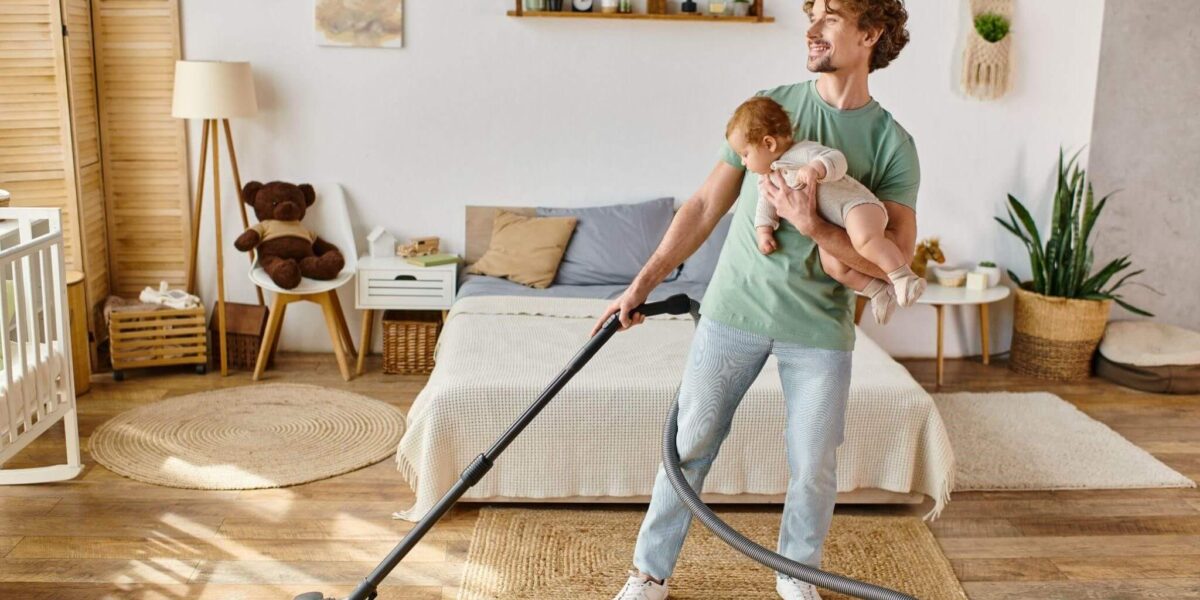 Ein Mann hält ein Baby auf einem Arm, während er in einem hellen, aufgeräumten Schlafzimmer mit hölzernen und gewebten Dekorationselementen den Boden saugt.
