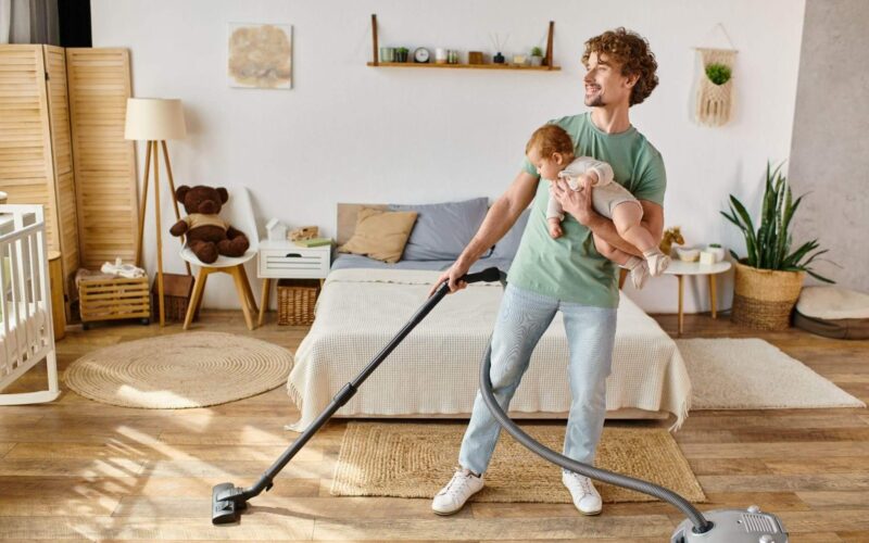 Ein Mann hält ein Baby auf einem Arm, während er in einem hellen, aufgeräumten Schlafzimmer mit hölzernen und gewebten Dekorationselementen den Boden saugt.