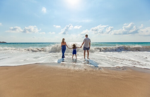 Drei Personen, zwei Erwachsene und ein Kind, stehen händchenhaltend am Rande des Ozeans an einem Sandstrand unter einem blauen Himmel mit vereinzelten Wolken.
