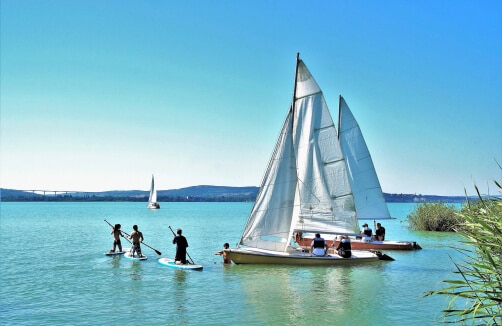 Menschen segeln und paddeln auf einem ruhigen See unter einem klaren blauen Himmel, mit entfernten Hügeln und einem weiteren Segelboot im Hintergrund.